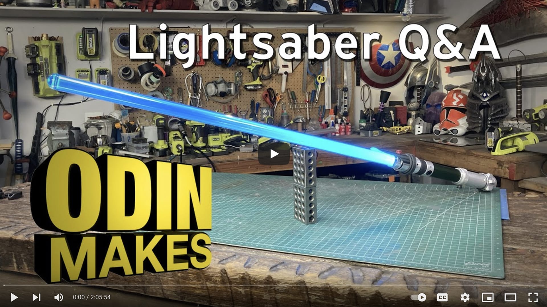 A workbench displays a lightsaber prop with the text "Lightsaber Q&A" at the top and "Odin Makes" at the bottom. Workshop tools and equipment are visible in the background, perfect for those eager to learn how Odin Abbott tackles projects like making a combat-ready lightsaber.