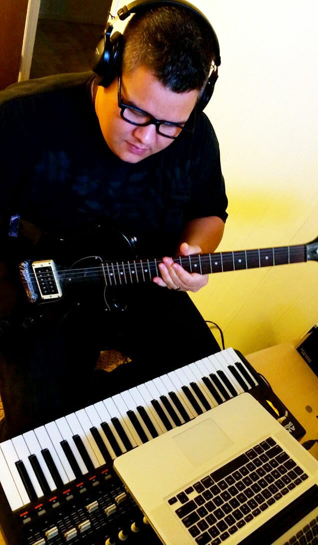 A man is playing a guitar in front of a laptop.