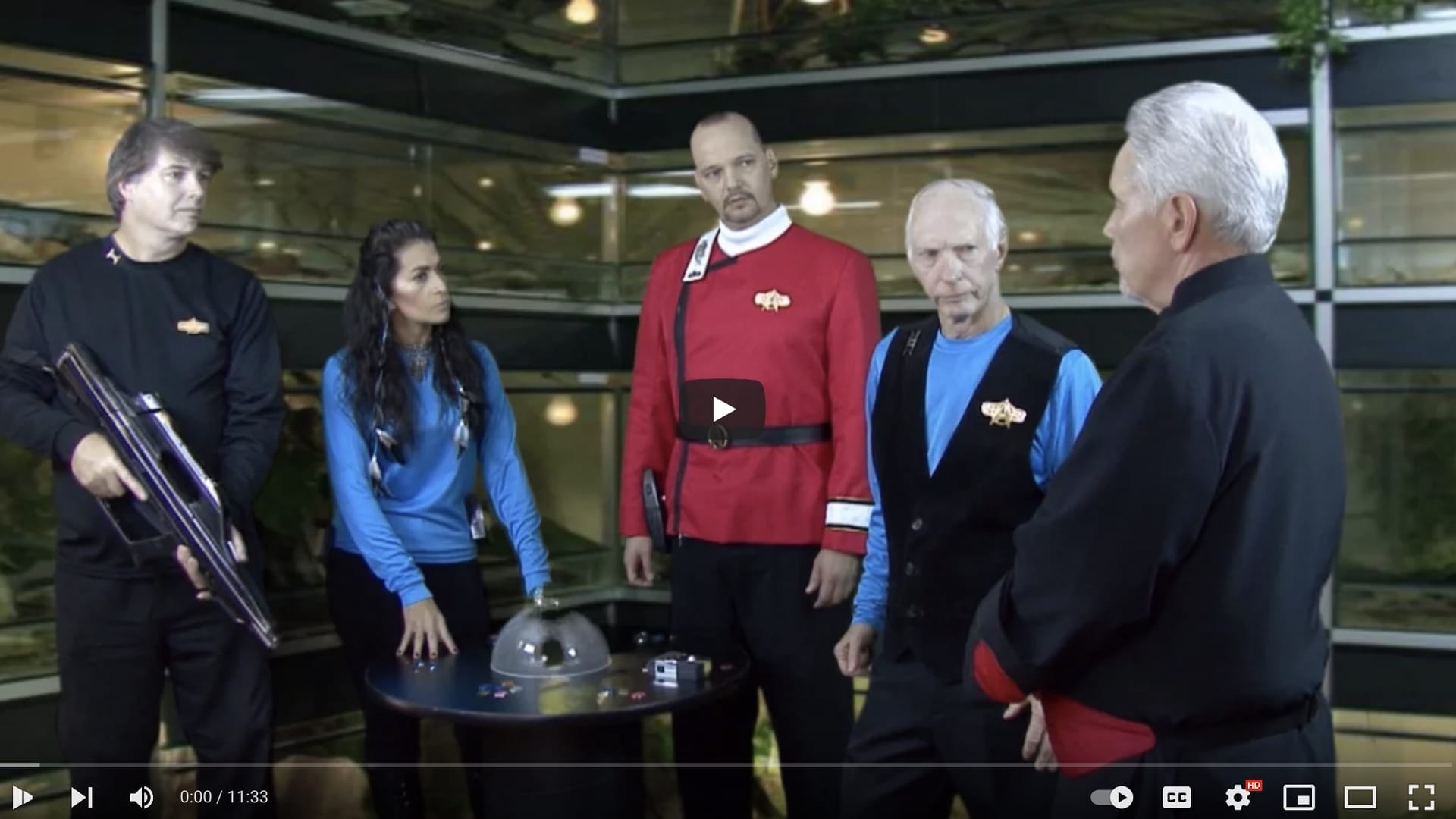 Five people in Star Trek uniforms stand in a futuristic room with a round table in the center. One person holds a large device, while others appear to be in conversation. A play button overlays the image, hinting at a new episode from Project Potemkin by Potemkin Pictures, perfect for Beyond Geek fans.